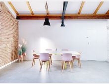 Minimalistischer Besprechungsraum mit einer weißen Wand, einem Tisch mit pastellrosa Stühlen und einer dekorativen Ziegelwand links. Die Decke zeigt sichtbare Holzbalken und schwarze Hängeleuchten, der Boden besteht aus Beton.