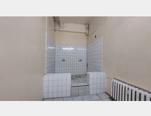 Ein einfacher Raum mit beigen Wänden, in dessen Zentrum eine kleine, geflieste Dusch- oder Waschstelle mit weißem Kachelmuster und zwei Wasserhähnen zu sehen ist; ein Heizkörper befindet sich an der rechten Wand.