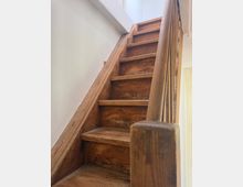 Schmale Holztreppe in einem Innenraum, mit abgenutzten Stufen und einem Holzgeländer an der rechten Seite. Weiß gestrichene Wände und ein helles Fenster bzw. Lichtöffnung oben am Treppenlauf.