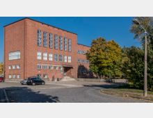 Backstein-Gebäude im Stil der modernen Architektur mit mehreren rechteckigen Fenstern, einem zentralen Eingangsbereich mit Treppen und einer gepflasterten Vorfläche. Rechts neben dem Gebäude befinden sich Bäume und ein Laternenmast.