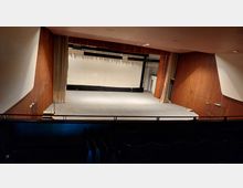 Leerer Theatersaal mit Blick auf eine Bühne. Die Bühne ist von holzverkleideten Wänden umgeben, mit beigen Vorhängen an den Seiten und einer weißen Rückwand. Im Vordergrund befinden sich leere Sitzreihen.