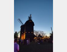 Holzverkleidete Windmühle mit seitlichen Flügeln und kleinem Aufbau an der Spitze, aufgenommen in der Dämmerung vor blauem Himmel. Im Vordergrund eine Wiese mit Lichterketten zwischen Bäumen und einem überdachten Sitzbereich.