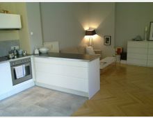 Moderner Wohnraum mit offener Küche und Wohnzimmerbereich. Die Küche hat weiße Schränke, eine schwarze Arbeitsfläche und einen eingebauten Ofen, das Wohnzimmer beinhaltet ein Sofa, einen Couchtisch, eine Stehlampe und eine Kommode auf einem hellen Parkettboden.