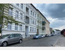 Straßenszene mit mehrstöckigen Wohnhäusern aus der Gründerzeit, verziert mit Stuckfassaden in Weiß, Grün und Beige. Auf der gepflasterten Straße parken mehrere Autos, und der Himmel ist bewölkt.