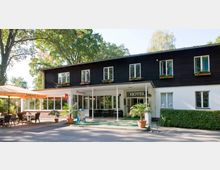 Zweistöckiges Hotelgebäude in einer grünen, bewaldeten Umgebung, mit einer dunklen Holzfassade im oberen Bereich und weißen Wänden im Erdgeschoss. Vor dem Eingang befinden sich ein mit Pflanzen dekorierter Eingangsbereich, ein beschatteter Außenbereich mit Tischen und Stühlen sowie ein Schild mit der Aufschrift "Rezeption".