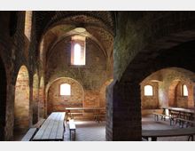 Innenansicht eines historischen Gewölberaums aus Ziegelstein mit romanischen Bögen, kleinen Fenstern und Kreuzrippengewölben. Im Raum stehen lange Holztische und Bänke auf einem Boden aus roten Fliesen.