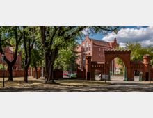 Historischer roter Backsteinbau mit Torbögen und Zinnen, umgeben von Bäumen in einem parkähnlichen Gelände. Im Hintergrund sind weitere Gebäude aus rotem Backstein sichtbar.