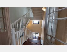 Treppenhaus in einem Altbau mit weiß gestrichenen Holzgeländern und braunen Holzstufen. Ein Fenster über den Treppen sorgt für Tageslicht, die Wände sind cremefarben gestrichen, und rechts befindet sich eine Tür mit gläsernen Sprossen.