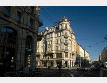 Straßenszene in einer Stadt mit einem historischen, vierstöckigen Eckgebäude mit Kuppeldach und verzierter Fassade. Im Vordergrund Straßenbahnschienen, geparkte Fahrräder und mehrere Geschäfte, darunter ein Café.