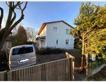 Zweistöckiges weißes Wohnhaus mit Satteldach, umgeben von Bäumen und Sträuchern. Im Vordergrund ein Holzzaun, dahinter ein geparkter silberner Van und ein kleiner Vorgarten.