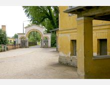 Ein sandiger Weg führt durch einen gemauerten Torbogen mit seitlichen Holztüren. Rechts ist ein gelbes, verputztes Gebäude mit verwitterten Wänden und Fenstern zu sehen, im Hintergrund stehen Bäume und weitere Gebäude.