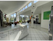 Moderne, helle Wohnküche mit offener Gestaltung, weißen Möbeln und großen Fensterflächen, die viel Tageslicht hereinlassen; im Hintergrund ein Wohnzimmerbereich mit Kamin, bunten Wandbildern und Blick ins Grüne.