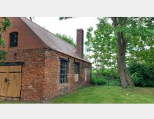 Backsteingebäude mit Satteldach, hölzernen Doppeltüren und einem hohen Schornstein, umgeben von Gras und Bäumen in einer ländlichen Umgebung.