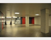 Unterirdischer Durchgang mit gefliesten Wänden in Weiß und Dunkelblau, roten Türen und hellem Deckenlicht. Rechts befinden sich weiße Säulen, und auf der linken Seite führen Rolltreppen und eine Treppe nach unten.