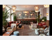 Ein stilvoll gestalteter Lounge-Bereich mit einer Mischung aus modernen und klassischen Möbeln, darunter bunte Sessel und Sofas sowie dekorative Stehlampen. Große Pflanzen, eine Wand mit abstrakter Kunst und warme Beleuchtung prägen das Ambiente, das auf einem dunklen Holzboden arrangiert ist.