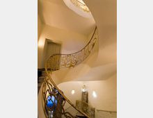 Geschwungene Wendeltreppe mit kunstvoll verziertem, goldfarbenem Geländer in einem Innenraum mit hellen Wänden. Im Hintergrund sind eine Tür, Wandlampen und eine Statue erkennbar.