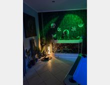 Innenraum eines Wellness- oder Meditationsbereichs mit gefliester Bodenfläche, grün beleuchteter Wand mit Blattmuster und dekorativen Symbolen. Vor der Wand stehen ein großes Terrarium oder Aquarium mit Buddha-Figur und Pflanzen sowie weitere Buddha-Dekorationen, Pflanzen und eine Stehlampe; rechts ist der Rand eines beleuchteten Whirlpools zu sehen.