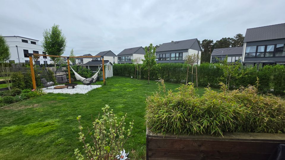 Blick über einen gepflegten Garten mit großer Rasenfläche, Hecken und jungen Bäumen in einem Wohngebiet mit modernen Einfamilienhäusern im Hintergrund. Links steht eine Holzpergola mit Hängesesseln und Hängematte auf einer Kiesfläche neben einem kleinen Gartenhaus.