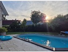 Gartenansicht mit rechteckigem Swimmingpool, umgeben von einer gefliesten Terrasse und Rasenfläche; im Hintergrund sind Hecken, Bäume und Wohnhäuser mit Satteldächern sowie ein Sonnenuntergang zu sehen.