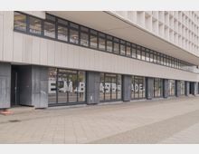 Moderne Fassade eines Gebäudes mit großflächigen Glasfenstern im Erdgeschoss, hinter denen der Schriftzug "EMBASSADE" sichtbar ist. Darüber befindet sich eine Reihe von Fenstern, die unter einem weit überstehenden weißen Vordach angeordnet sind.