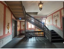 Treppenhaus in einem historischen Gebäude mit dekorativen Wänden in Rot- und Beigetönen, verziert mit gemalten Vasenmotiven. Eine Holztreppe mit ornamentalem Geländer führt nach oben, beleuchtet von einem dekorativen Kronleuchter; ein Fenster sorgt für zusätzliches Tageslicht.
