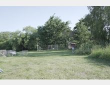 Grüne Wiese mit Bäumen und einem kleinen Zaun im Hintergrund. Rechts befinden sich eine Holzhütte mit Wimpelgirlande sowie Hühner und eine Liege im Gras.