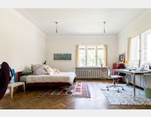 Ein geräumiges Schlafzimmer mit einem Arbeitsbereich. Links steht ein Bett mit mehreren Kissen auf bunten Teppichen, rechts ein Schreibtisch mit Bürostuhl und Computer, daneben Fenster mit beigefarbenen Vorhängen, die Tageslicht hereinlassen.