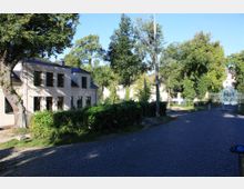 Wohngebiet mit einem zweistöckigen, hellen Gebäude mit schwarzen Fenstern und einem schrägen Dach rechts im Vordergrund. Im Hintergrund befinden sich Bäume, weitere Gebäude und ein schmiedeeisernes Tor; der Boden ist mit Kopfsteinpflaster bedeckt.