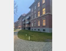 Mehrstöckiges Wohngebäude aus Backstein mit großen Fenstern, teils mit eingeschaltetem Innenlicht, sowie einigen Balkonen. Davor befinden sich ein gepflasterter Gehweg, eine Rasenfläche und vereinzelt Laternenpfähle; die Szene spielt bei Dämmerung.