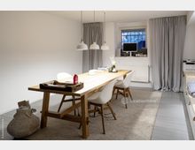 Ein modern eingerichteter Raum mit einem großen Holztisch, umgeben von weißen Stühlen auf einem hellen Teppich. An der Wand befindet sich ein Fenster mit grauen Vorhängen, daneben steht ein Heizkörper, und auf dem Tisch liegen Dekorationsgegenstände sowie Arbeitsmaterialien.