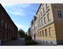 Eine ruhige Straße mit historischen Gebäuden, links ein rotes Backsteingebäude und rechts eine Reihe von gelb und pastellfarben verputzten Wohnhäusern. In der Mitte der Straße befinden sich Bäume und eine gepflasterte Gehfläche.