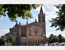 Backsteinkirche mit neoromanischer Architektur, einschließend großen Rundbogenfenstern, einem zentralen Rosettenfenster und einem hohen Glockenturm mit Spitzdach; umgeben von Bäumen und parkenden Fahrzeugen im Vordergrund.