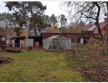 Ein Garten mit einem kleinen Gewächshaus im Vordergrund, umgeben von kahlen Bäumen und Sträuchern. Im Hintergrund stehen eine Holzhütte und weitere Gebäude mit roten und dunklen Dächern, eingebettet in eine waldreiche Umgebung.