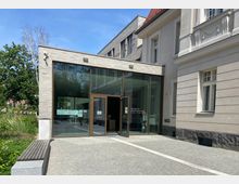 Eingangsbereich eines Amtsgerichts mit moderner Glasfassade und angrenzendem historischen Gebäude mit Putzfassade. Der Vorplatz ist gepflastert und von grüner Bepflanzung umgeben.