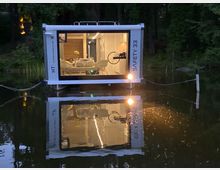 Ein modernes Hausboot mit großer Fensterfront, das auf einem ruhigen Waldsee schwimmt. Das Innere ist hell beleuchtet und zeigt einen Essbereich mit Tisch und Stühlen; die Umgebung ist von Bäumen umgeben, und das Boot ist mit Seilen befestigt.