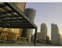 Ansicht des Potsdamer Platzes in Berlin mit moderner Architektur, einschließlich hoher Glas- und Bürogebäude. Im Vordergrund ein überdachter Eingang mit der Aufschrift "Potsdamer Platz" und das Bahnhofsgebäude mit DB-Logo.