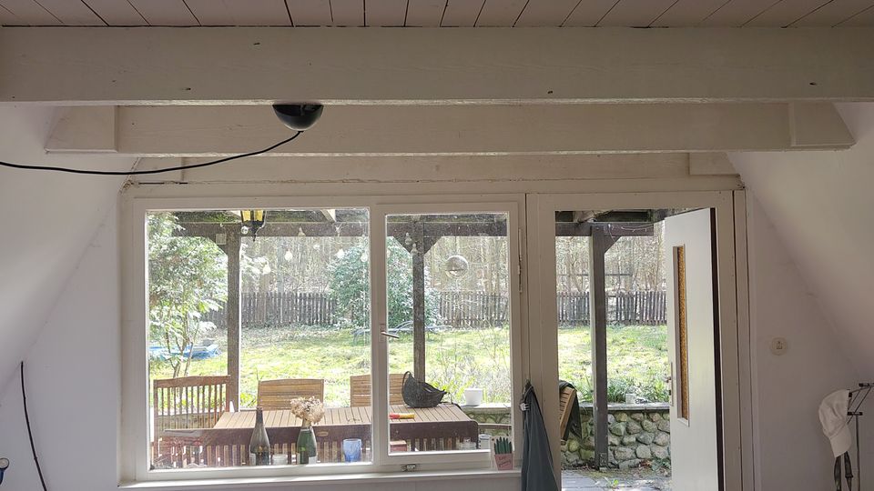 Blick aus einem Wohnraum mit sichtbaren weißen Deckenbalken auf ein großes Fenster und eine geöffnete Tür zur Terrasse. Draußen sind eine überdachte Sitzgruppe, Steinplatten und ein grüner Garten mit Holzzaun zu sehen.