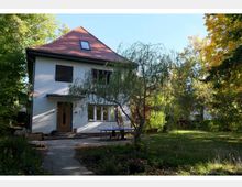 Zweistöckiges Einfamilienhaus mit weißer Fassade und rotem Satteldach, umgeben von einem großen Garten mit Bäumen und Sträuchern. Ein gepflasterter Weg führt zur Haustür, neben der ein Tisch im Freien steht.