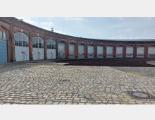 Ein historisches, halbrundes Backsteingebäude mit großen Toren, vermutlich ein Lokschuppen. Im Vordergrund sind ein gepflasterter Hof und mehrere Schienenstränge sichtbar, die in das Gebäude führen.