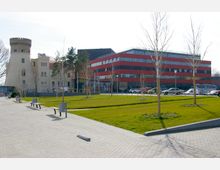 Moderne Gebäudekombination mit einem historischen, beigen Turm auf der linken Seite und einem roten, mehrstöckigen Bürogebäude rechts. Davor ein gepflasterter Weg, Rasenflächen und dünne, junge Bäume, mit parkenden Autos im Hintergrund.