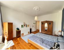 Schlafzimmer mit Holzboden und weißen Wänden. Der Raum verfügt über ein Doppelbett mit grauer Bettwäsche, einen großen Holzkleiderschrank mit Spiegeleinsatz, einen kleineren Holzschrank, ein weißes Regal sowie eine Deckenleuchte mit Stuckverzierung.