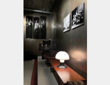 Ein stilvoller Innenraum mit dunklen Wänden und moderner, gedämpfter Beleuchtung. An der Wand hängen schwarz-weiße Fotografien, darunter Portraits und Blumenmotive, während im Vordergrund ein langer Holztisch mit einer auffälligen Pilzlampe und lederbezogene Stühle zu sehen sind.