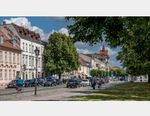 Straßenszene in einer kleinen Stadt mit historischen Gebäuden, darunter ein Geschäft mit der Aufschrift „Juwelier Hallex“, und einer Reihe parkender Autos. Im Vordergrund eine Radfahrerin auf einem Fahrradweg, Fußgänger, und eine baumbestandene Grünfläche auf der rechten Seite.