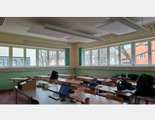 Klassenzimmer mit mehreren Reihen aus Holztischen und Stühlen sowie einem Lehrerpult im Vordergrund. Große Fensterfronten mit Blick auf Bäume und umliegende Gebäude, grün- und beigefarbene Wände und abgehängte Deckenpaneele mit Leuchtstofflampen prägen den Raum.