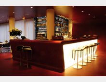 Moderne Bar mit geschwungenem, beleuchtetem Tresen aus weißem Material und roten Wänden. Im Hintergrund befindet sich ein Regal mit einer Auswahl an Spirituosen und Gläsern, daneben stehen mehrere Barhocker mit schwarzem Sitz.