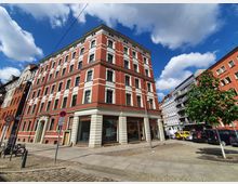 Eckgebäude im Stil der Gründerzeit mit roter Klinkerfassade, weißen Akzenten und großen Fenstern, gelegen an einer gepflasterten Straße mit Gehwegen. Umgeben von weiteren mehrstöckigen Wohnhäusern und parkenden Autos, unter einem Himmel mit vereinzelten Wolken.