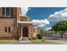 Teilansicht einer historischen Ziegelkirche mit runden und rechteckigen Fenstern sowie einem grünen Kupferdach, umgeben von einer Straße, Grünflächen und Bäumen. Im Hintergrund ist modernes Stadtbild mit Gebäuden und blauen Himmel mit Wolken zu sehen.