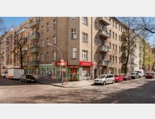 Ecke einer Wohnstraße in einer Stadt mit einem mehrstöckigen, verputzten Altbau. Im Erdgeschoss befinden sich Geschäfte mit roten Markisen, in der Umgebung sind parkende Autos, Kopfsteinpflaster und vereinzelte Bäume sichtbar.