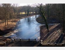 Gefrorener Teich in einem Park, umgeben von kahlen Bäumen und einem mit Laub bedeckten Boden, mit einer steinernen Balustrade im Vordergrund. Im Hintergrund ist eine offene Wiesenfläche sichtbar.