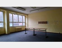 Leerer, älterer Büroraum mit gelb gestrichenen Wänden, Teppichboden und einer langen Tischreihe in der Mitte. Große Fensterfront mit Heizkörper darunter und Deckenleuchten; an der rechten Wand sind entfernte Verkleidungen bzw. abgeblätterte Stellen sichtbar.